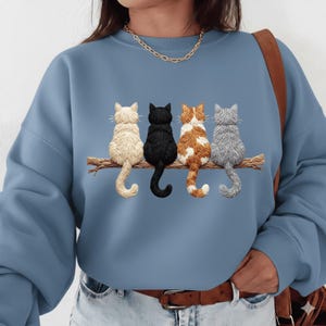 Könnte beinhalten: Hellblaues Sweatshirt mit einem gestickten Design von vier Katzen, die auf einem Ast sitzen. Die Katzen sind in den Farben Creme, Schwarz, Orange und Weiß sowie Grau gehalten. Das Sweatshirt hat lange Ärmel und einen Rundhalsausschnitt.