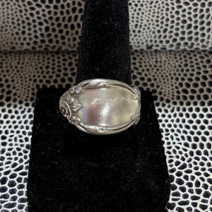 May include: A silver ring with an oval face, displayed on a black velvet stand. The ring has decorative floral and scrollwork around the edges of the face. The ring's face is smooth and reflective, catching the light.