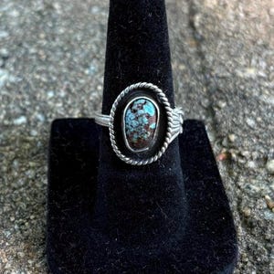 May include: A silver ring with an oval turquoise stone. The stone is set in a silver bezel with a twisted rope design. The ring is displayed on a black velvet ring holder.