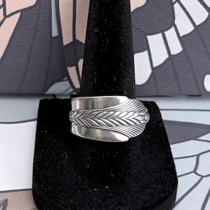 May include: A silver-toned spoon ring featuring an embossed wheat stalk pattern that wraps around the band. The metal has a polished finish with detailed, textured lines carved into the surface of the wheat design, creating a distinct botanical motif.