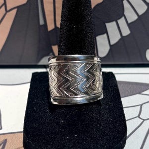 May include: A wide silver-toned metal ring featuring a repeating embossed chevron pattern across the center band. The ring has smooth, polished edges at the top and bottom, creating a distinct contrast with the textured, geometric zigzag design.