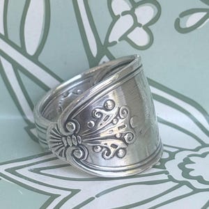 May include: A silver-toned ring crafted from a repurposed spoon handle, featuring an embossed decorative pattern with curved lines, small circular accents, and a fan-like motif at the base, showing a polished metallic finish and intricate relief.