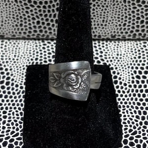 Könnte beinhalten: Ein silberner Ring mit rechteckiger Oberfläche, der ein erhabenes Rosendesign aufweist. Der Ring wird auf einem schwarzen Samtringhalter vor einem schwarz-weiß gemusterten Hintergrund präsentiert. Der Ring scheint aus recyceltem Besteck hergestellt zu sein.