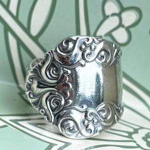 May include: A silver spoon ring featuring an intricate embossed scrollwork pattern along the handle section. The metal surface is polished and reflective, showcasing detailed swirling motifs and small raised dots integrated into the design.