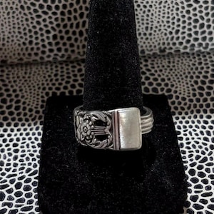 Puede incluir: Un anillo plateado con una piedra rectangular de color claro y detalles florales ornamentados. El anillo se exhibe en un soporte de anillo de terciopelo negro sobre un fondo estampado.