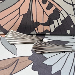 Puede incluir: Dos horquillas de metal plateado con un diseño en forma de abanico y estrías verticales. Las horquillas tienen un acabado pulido y reflectante, con una silueta cónica que se ensancha hacia el borde superior, creando una textura acanalada.