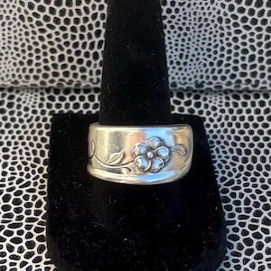 May include: A silver ring with a floral design. The ring features a raised flower with detailed petals and leaves. The band is wide and has a slightly textured surface. The ring is displayed on a black velvet ring holder.