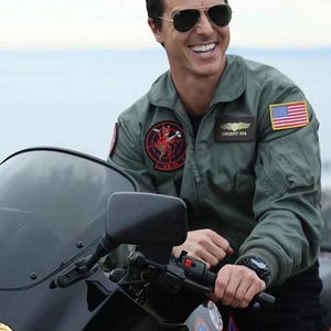 May include: A person wearing aviator sunglasses and a green bomber jacket with patches, including a US flag and the text "COCKPIT USA". The person is smiling and sitting on a black motorcycle.