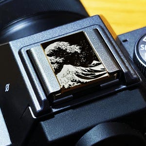 May include: A black camera hot shoe cover featuring a high-contrast white and black graphic print of a large ocean wave and a mountain peak. The square cover is inserted into the metal hot shoe mount on top of a black digital camera body.