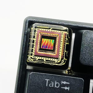 Peut inclure: Une touche de clavier personnalisée avec un motif carré multicolore encapsulé dans de la résine transparente. La touche noire est placée sur un clavier noir. Le mot "Tab" est visible sur la touche adjacente.