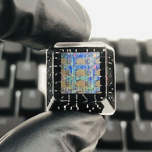 May include: A square, silver ring featuring a microchip design. The ring's face showcases a detailed, colourful microchip pattern. The ring is held by a gloved hand, with a keyboard in the background.