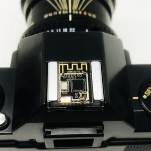 May include: Close-up of a black camera body with a lens attached. The top of the camera features a small circuit board with the text "LYR153 V1.0". A dial with "SELF PROGRAM" is visible. The camera is on a white surface.