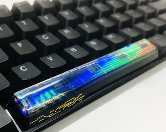 May include: Close-up of a black mechanical keyboard with a custom keycap. The keycap has a holographic design with blue, green, and yellow hues. The keyboard keys are black with white lettering.