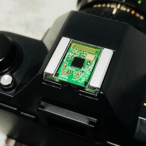 May include: Close-up of a black camera body with a circuit board mounted on top. The circuit board is green with a complex pattern of lines and components. The camera has a lens and various dials.