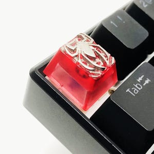 Könnte beinhalten: Eine rote, transparente Tastenkappe mit einem silbernen Spinnenmuster, die auf einer schwarzen Tastatur angebracht ist. Die Tastenkappe ist ein individuelles Design, wahrscheinlich für eine mechanische Tastatur.