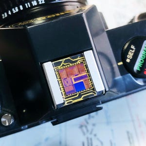 May include: Close-up of a black camera body with a lens and a small, colorful circuit board. The circuit board has a gold border and features shades of blue, purple, and brown. The camera's lens has aperture settings.