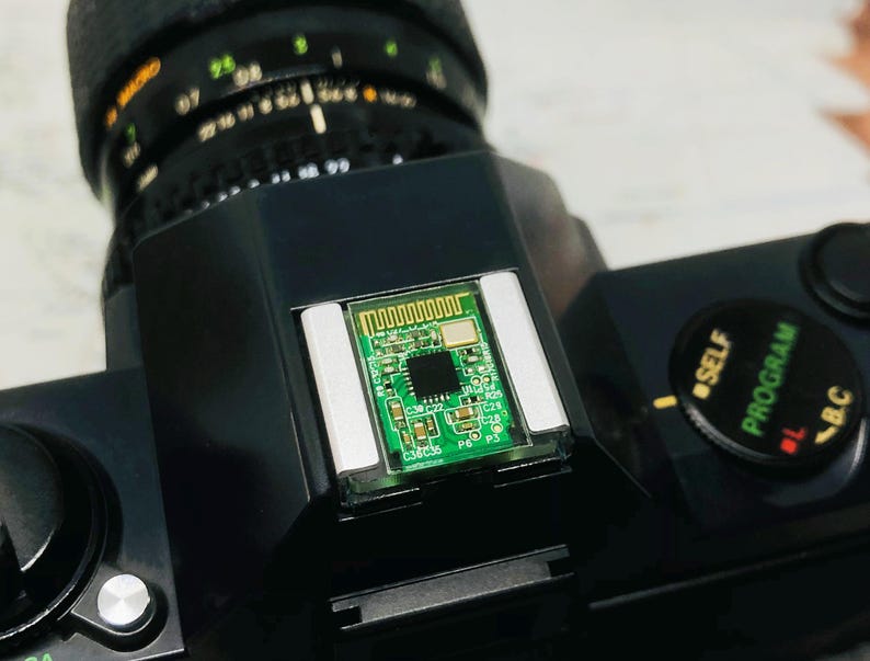 May include: Close-up of a black camera body with a lens attached. A small green circuit board is mounted on top of the camera. The lens has markings in white and orange. The camera has a dial with the words "SELF", "PROGRAM", and "B.C."