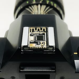 May include: Close-up of a black camera body with a lens attached. A small circuit board with the text "LY88153 V1.0" is mounted on the top. The camera has a self-timer dial and a silver-colored flash mount.