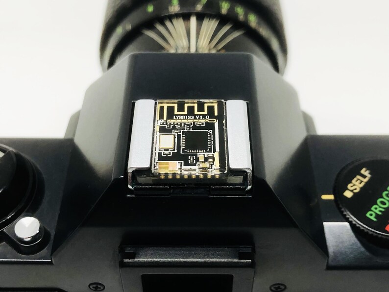 May include: Close-up of a black camera body with a lens attached. A small circuit board with the text "LY88153 V1.0" is mounted on the top. The camera has a self-timer dial and a silver-colored flash mount.