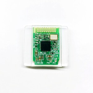 May include: A small, green circuit board with various electronic components, including a central black chip, is encased in a clear plastic holder. The board features several small, labeled components and a series of parallel lines at the top.