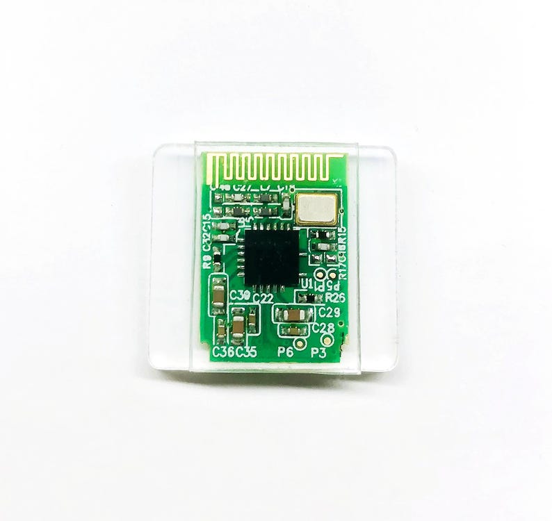 May include: A small, green circuit board with various electronic components, including a central black chip, is encased in a clear plastic holder. The board features several small, labeled components and a series of parallel lines at the top.