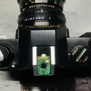 May include: Black Canon AE-1 camera with a lens attached. The camera has a silver lens and a small green circuit board in the center. The camera's settings include ASA, SELF, and PROGRAM.