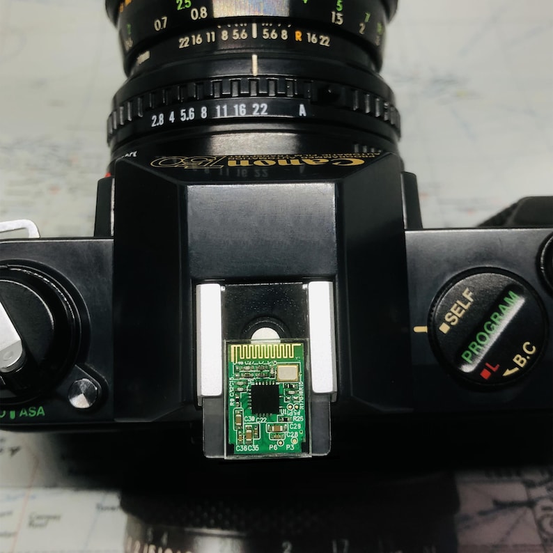May include: Black Canon AE-1 camera with a lens attached. The camera has a silver lens and a small green circuit board in the center. The camera's settings include ASA, SELF, and PROGRAM.
