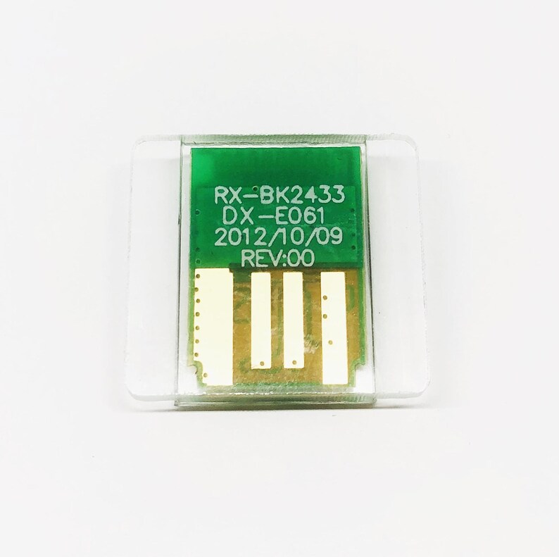 May include: A small, rectangular electronic component with a green circuit board and gold-colored connectors. The text "RX-BK2433 DX-E061 2012/10/09 REV:00" is printed on the green surface. The component is encased in a clear plastic housing.