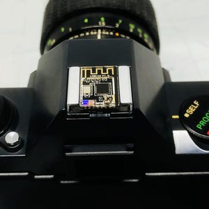 May include: Black camera body with a lens and a small circuit board attached to the top. The circuit board has the text "LYR153 V1.0" printed on it. The camera has a self-timer and program dial.