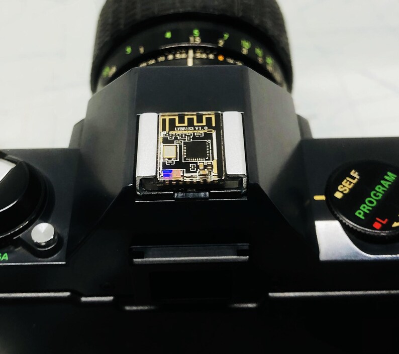 May include: Black camera body with a lens and a small circuit board attached to the top. The circuit board has the text "LYR153 V1.0" printed on it. The camera has a self-timer and program dial.