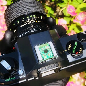 May include: A black and silver vintage camera with a Sigma lens. The lens has various settings, including f-stops and focal lengths. The camera body has an ISO/ASA dial and a program dial. A small circuit board is visible.