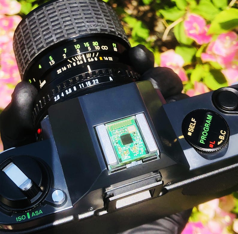 May include: A black and silver vintage camera with a Sigma lens. The lens has various settings, including f-stops and focal lengths. The camera body has an ISO/ASA dial and a program dial. A small circuit board is visible.