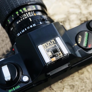 May include: A black vintage camera with a lens featuring aperture and focus settings. The camera body has buttons for self-timer and program settings. The top of the camera has a small circuit board.