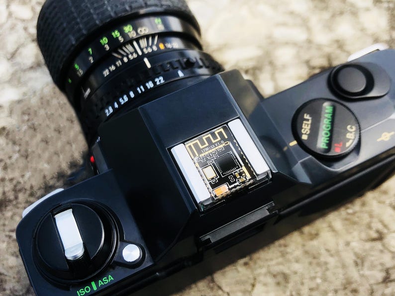 May include: A black vintage camera with a lens featuring aperture and focus settings. The camera body has buttons for self-timer and program settings. The top of the camera has a small circuit board.