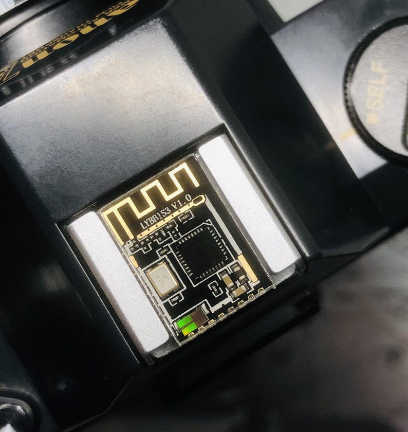May include: Close-up of a black electronic component with a gold-colored circuit board. The board has the text "LYBB!S3 V1.0" and a Wi-Fi symbol. The component is set in a black housing.