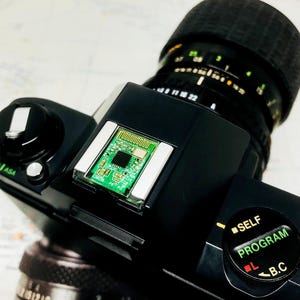May include: Close-up of a black camera with a lens and a green circuit board. The lens has measurement markings. The camera has a self-program dial and an ASA dial. The background is a map.