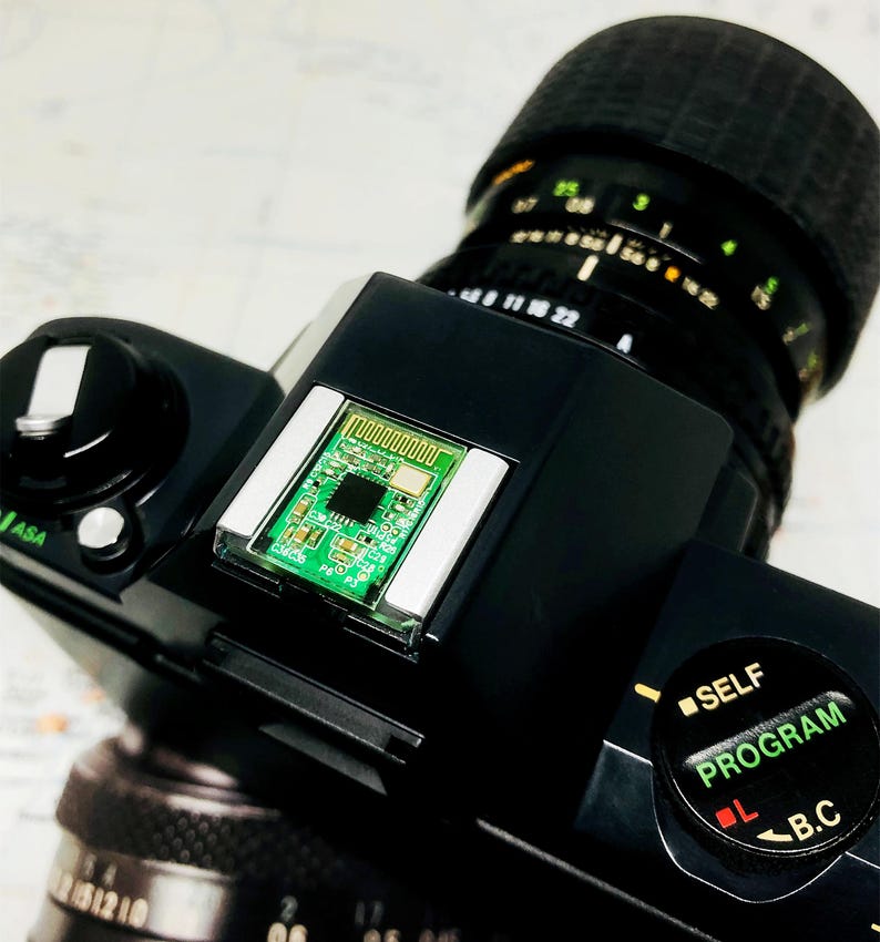 May include: Close-up of a black camera with a lens and a green circuit board. The lens has measurement markings. The camera has a self-program dial and an ASA dial. The background is a map.