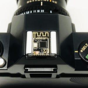 May include: Close-up of a black camera with a lens and a small circuit board labeled "LYMB153 V1.0." The camera has a dial with "SELF" and "PROGRAM" markings. The ASA setting is visible.