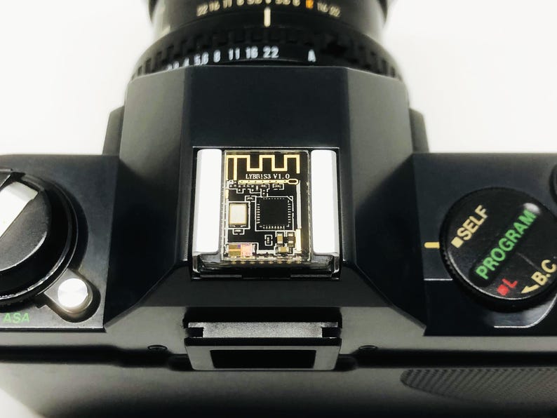 May include: Close-up of a black camera with a lens and a small circuit board labeled "LYMB153 V1.0." The camera has a dial with "SELF" and "PROGRAM" markings. The ASA setting is visible.