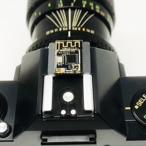 May include: Close-up of a black camera with a lens and a small circuit board attached to the top. The circuit board has the text "LYMBISS V1.0". The camera has a dial with the text "SELF PROGRAM".