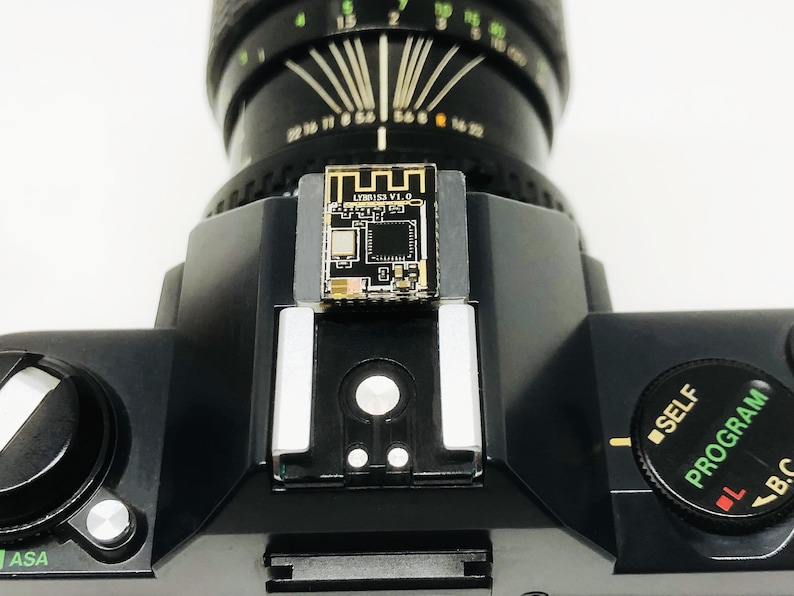 May include: Close-up of a black camera with a lens and a small circuit board attached to the top. The circuit board has the text "LYMBISS V1.0". The camera has a dial with the text "SELF PROGRAM".