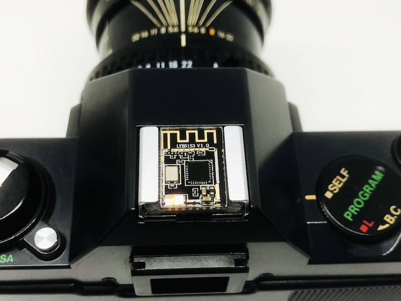 May include: Close-up of a black camera body with a lens attached. The top of the camera features a small circuit board with the text "LYR153 V1.0". A dial with "SELF PROGRAM" is visible. The camera is on a white surface.