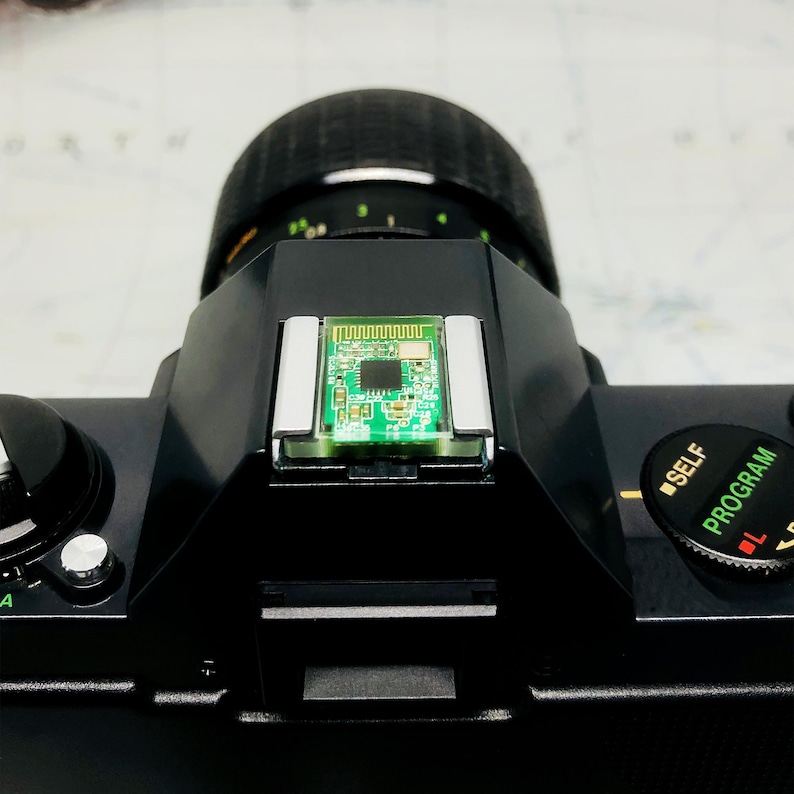 May include: Close-up of a black camera body with a lens and a small green circuit board. The camera has a dial with the words "SELF" and "PROGRAM" on it. The lens is black and has a ridged texture.