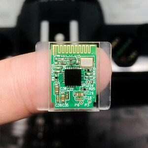 May include: A small, green circuit board with various electronic components, including a black square chip and a gold-colored antenna. The board is mounted on a clear plastic base.