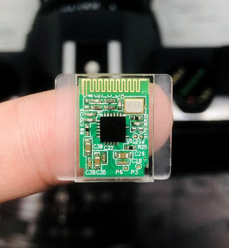 May include: A small, green circuit board with various electronic components, including a black square chip and a gold-colored antenna. The board is mounted on a clear plastic base.