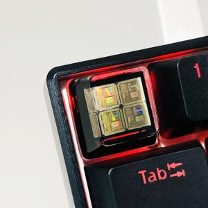 Puede incluir: Un teclado mecánico negro con retroiluminación roja. El teclado tiene una tecla personalizada con un diseño arcoíris iridiscente. La tecla está etiquetada como "Tab".