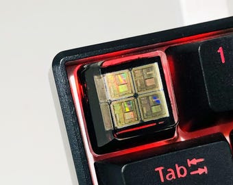 May include: A black mechanical keyboard with a red backlight. The keyboard features a custom keycap with a rainbow iridescent design. The keycap is labelled "Tab".