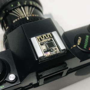May include: Black vintage camera with a silver and gold electronic component attached to the top. The component has the text "LYB81 V1.0" and a Wi-Fi symbol. The camera lens is black and the body has various dials and buttons.