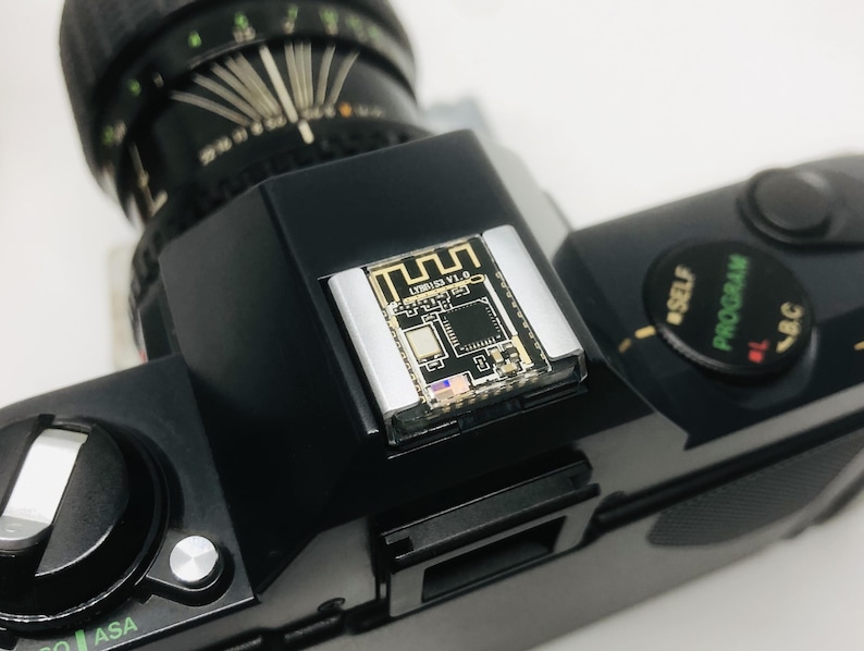 May include: Black vintage camera with a silver and gold electronic component attached to the top. The component has the text "LYB81 V1.0" and a Wi-Fi symbol. The camera lens is black and the body has various dials and buttons.