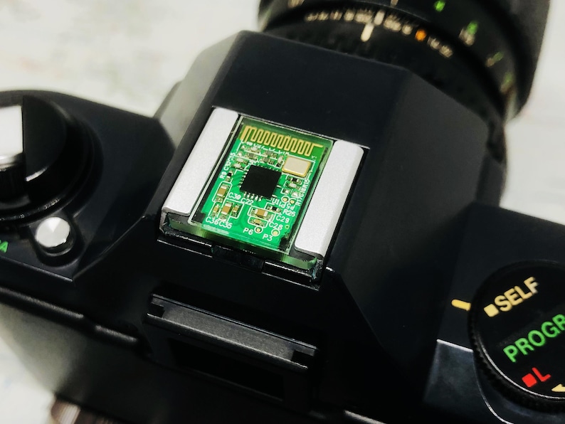 May include: Close-up of a black camera body with a circuit board mounted on top. The circuit board is green with a complex pattern of lines and components. The camera has a lens and various dials.